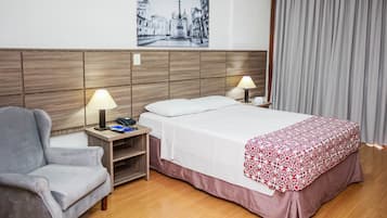 Quarto de hotel com uma cama, duas luminárias de cabeceira, uma cadeira, mesa de cabeceira, quadro na parede e janela com cortinas.