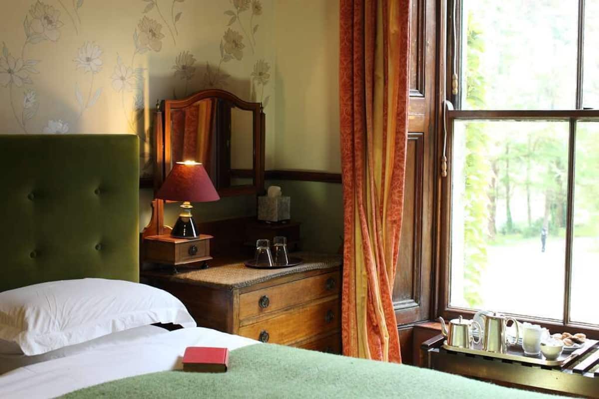 A bedroom with a green upholstered headboard, a wooden nightstand with a lamp, a dresser, and a window with curtains.
