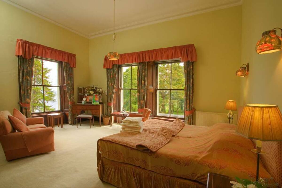 A bedroom with a large bed, a desk, a chair, and two windows with curtains.
