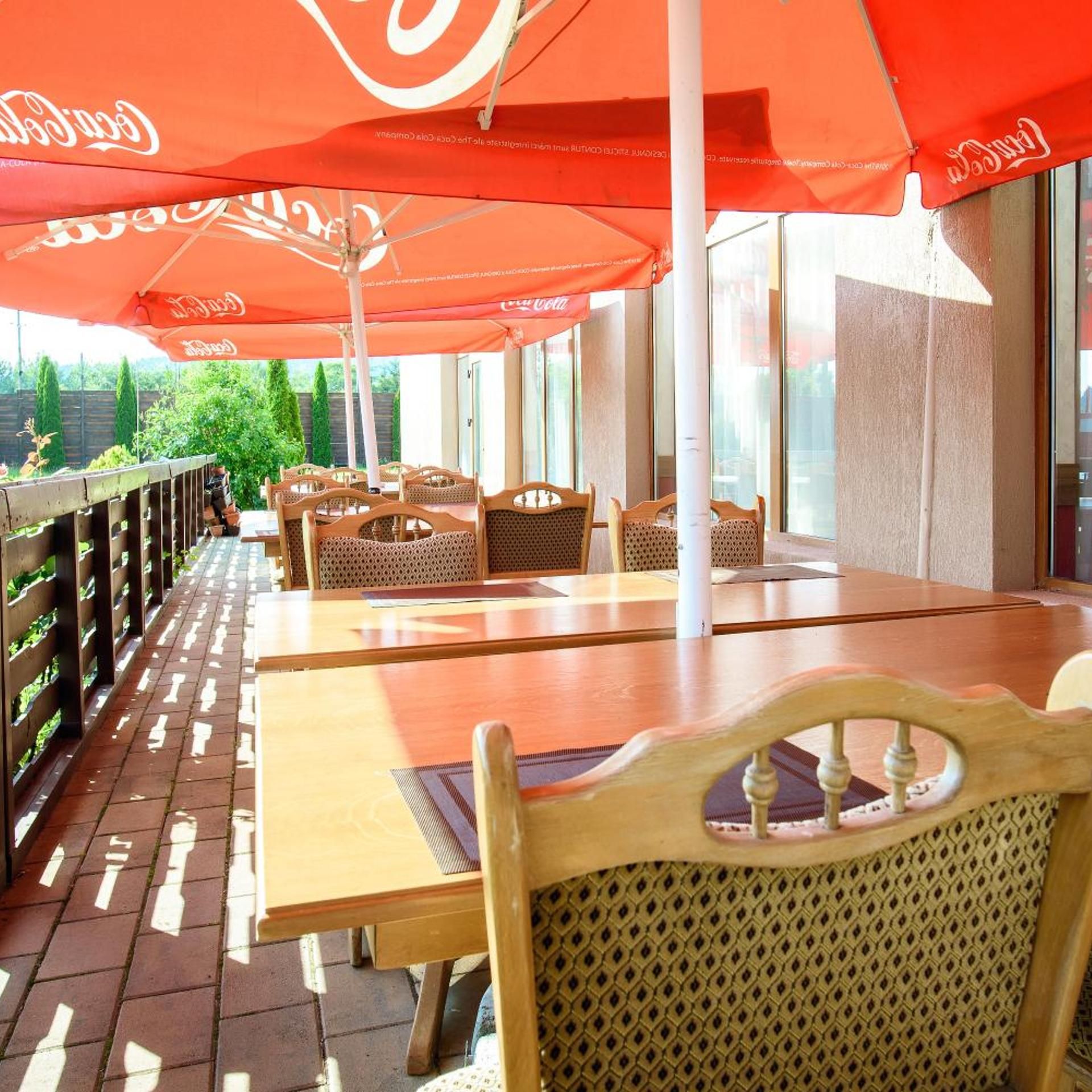 An outdoor dining area with wooden tables and chairs, red umbrellas, and a view of greenery.