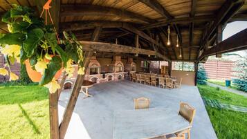 An outdoor patio area with brick barbecue grills, wooden tables and chairs, and hanging plants.