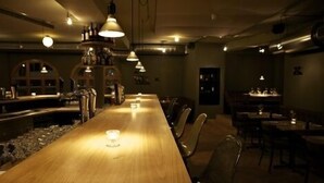 A dimly lit restaurant with a long wooden table and chairs, overhead lighting, and a bar area with shelves of bottles. - Arena Lodge (Flims)
