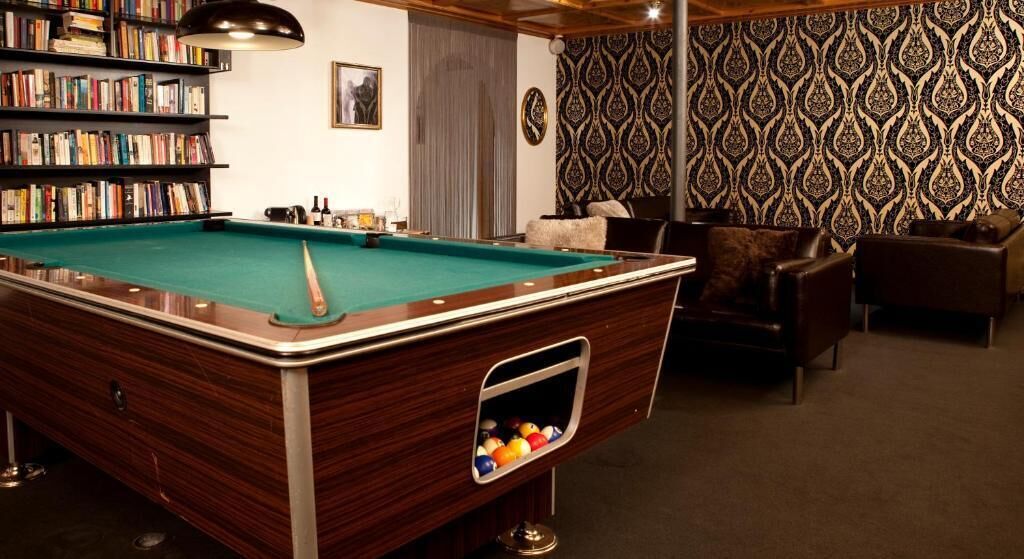 A pool table with a built-in ball rack, surrounded by leather chairs and a bookshelf filled with books.