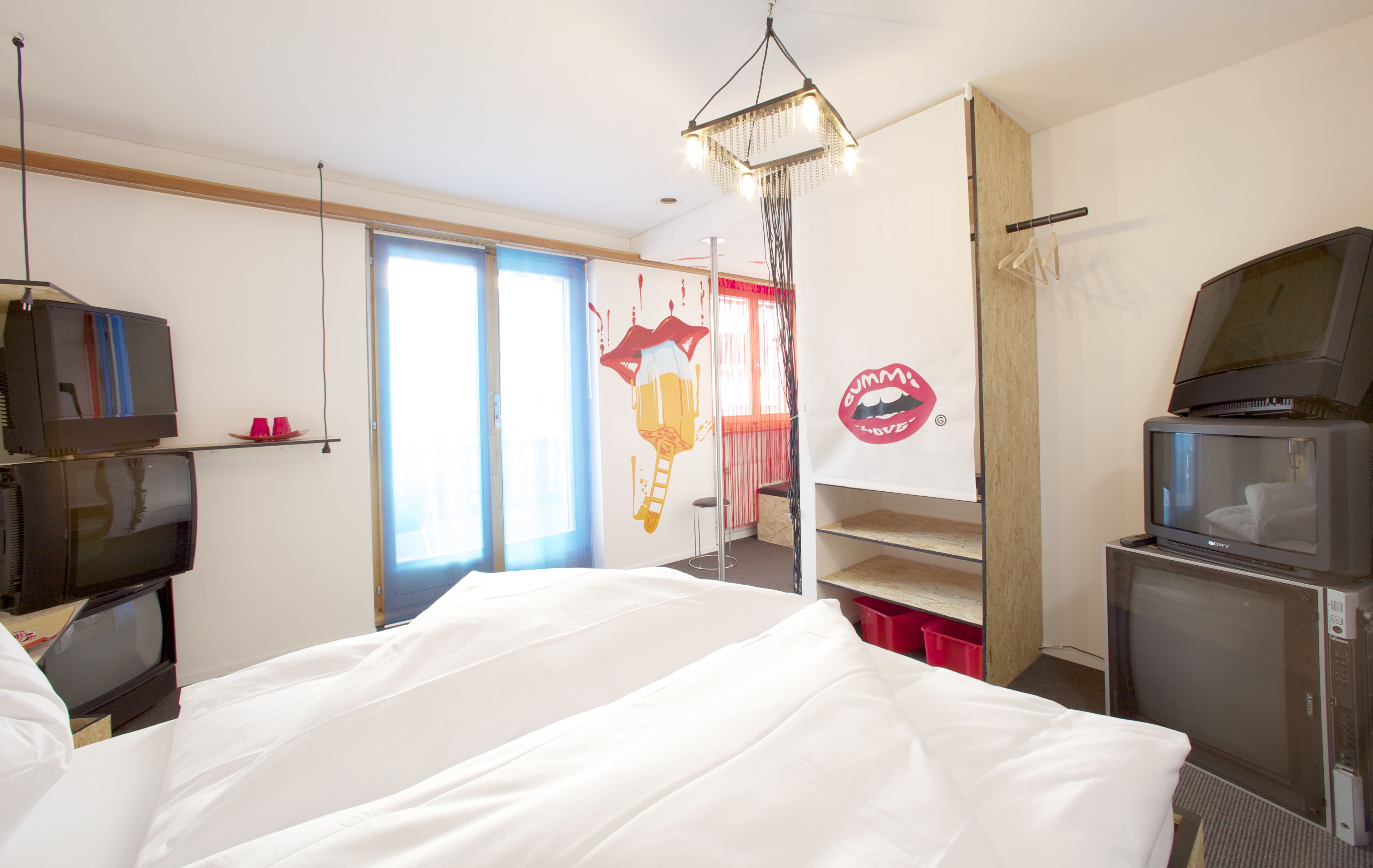 Ein Hotelzimmer mit einem Bett, zwei Fernsehern, einer Kristallleuchtre und einem roten Wandbild, sowie einem Fenster mit Vorhängen.