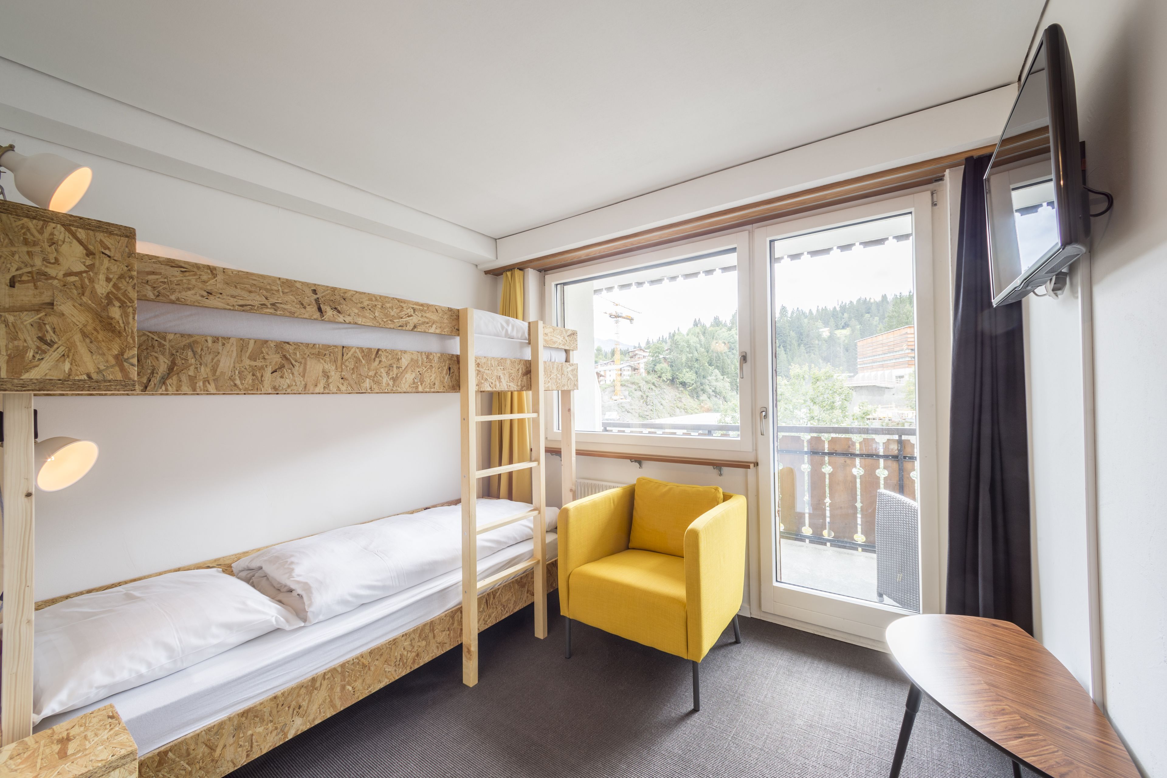 Una habitación con literas, un sillón amarillo, un televisor y una ventana con vistas a un balcón.