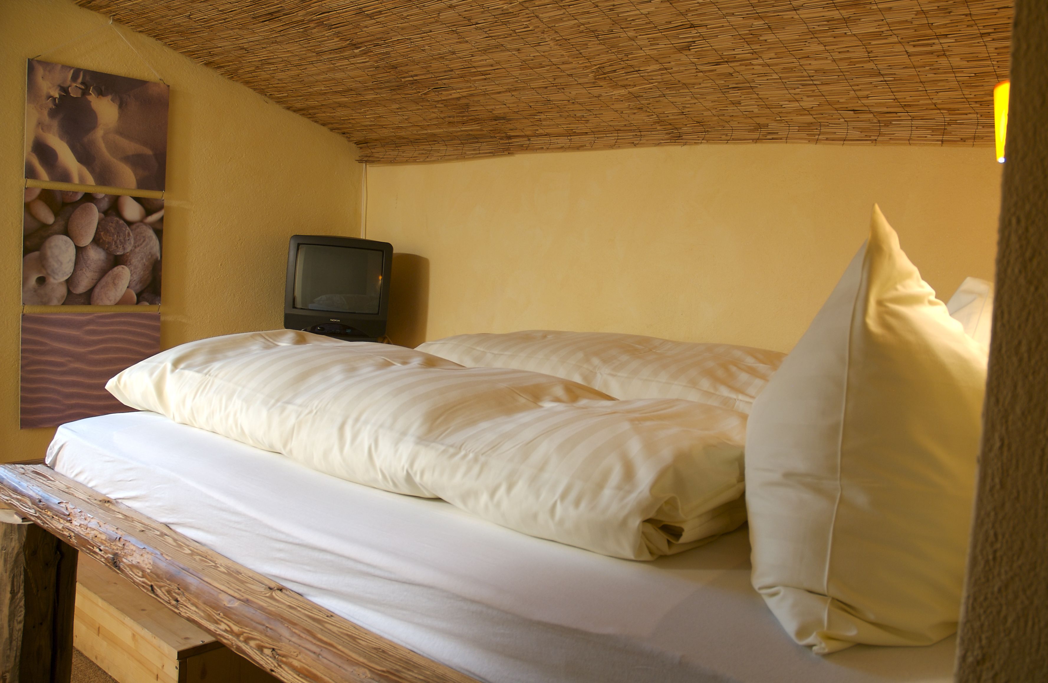 Una cama individual con ropa de cama blanca en una habitación con techo de paja y un televisor sobre un soporte de madera.