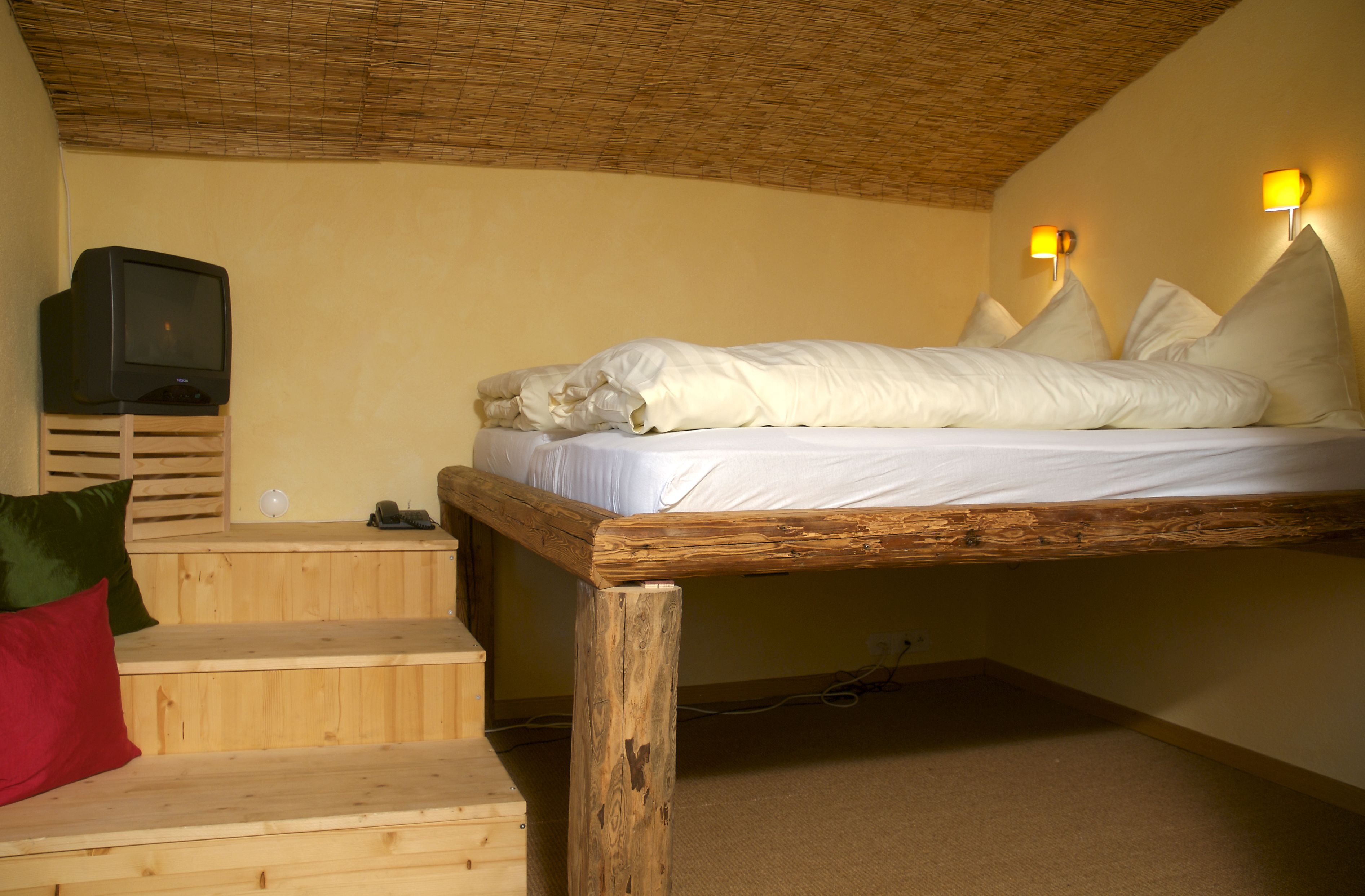 A single bed with white bedding, a television on a wooden stand, and a staircase with a red pillow.