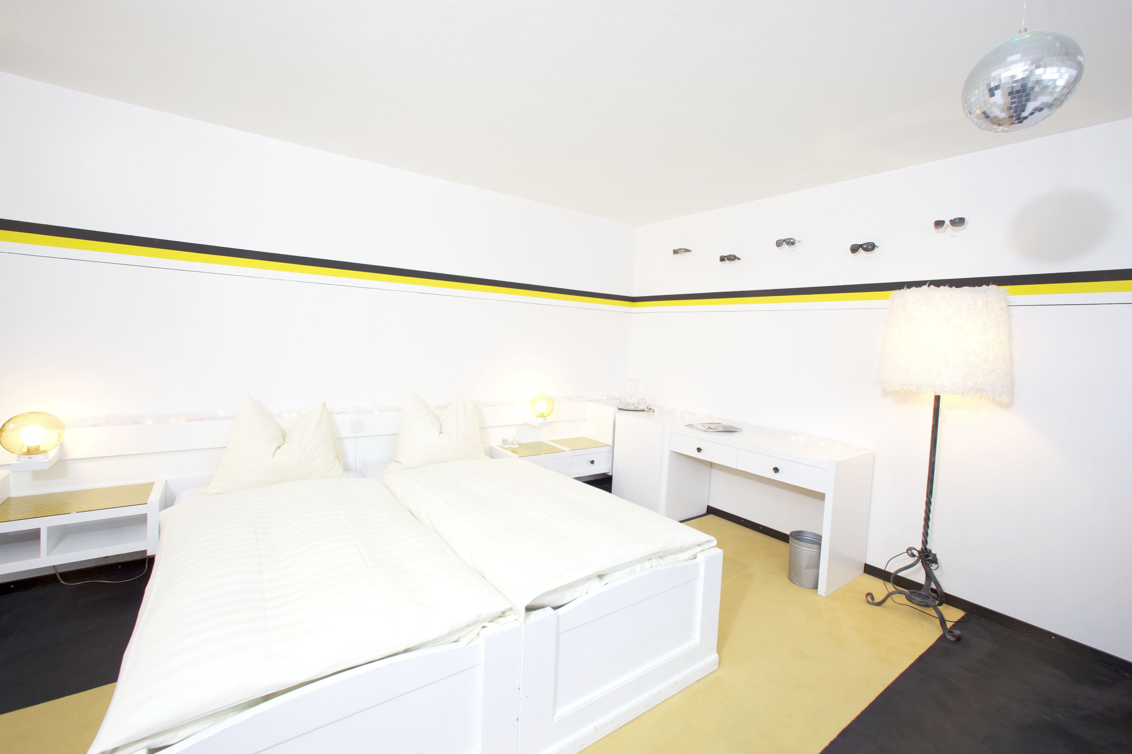 Et hotelværelse med seng, sengeborde, et skrivebord, en lampe og en skraldespand.