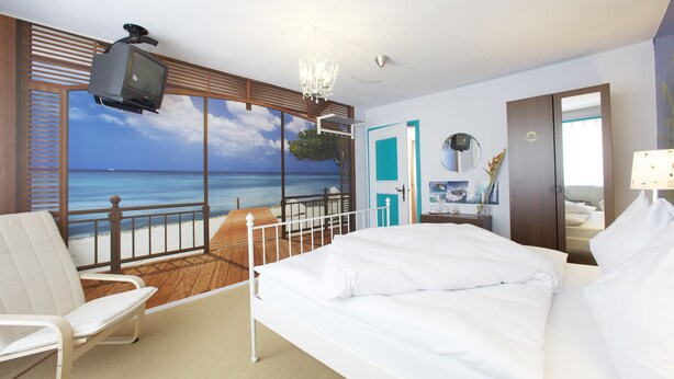A hotel room with a bed, a chair, a TV, a desk with a mirror, and a view of the ocean through a large window. - Arena Lodge (Flims)
