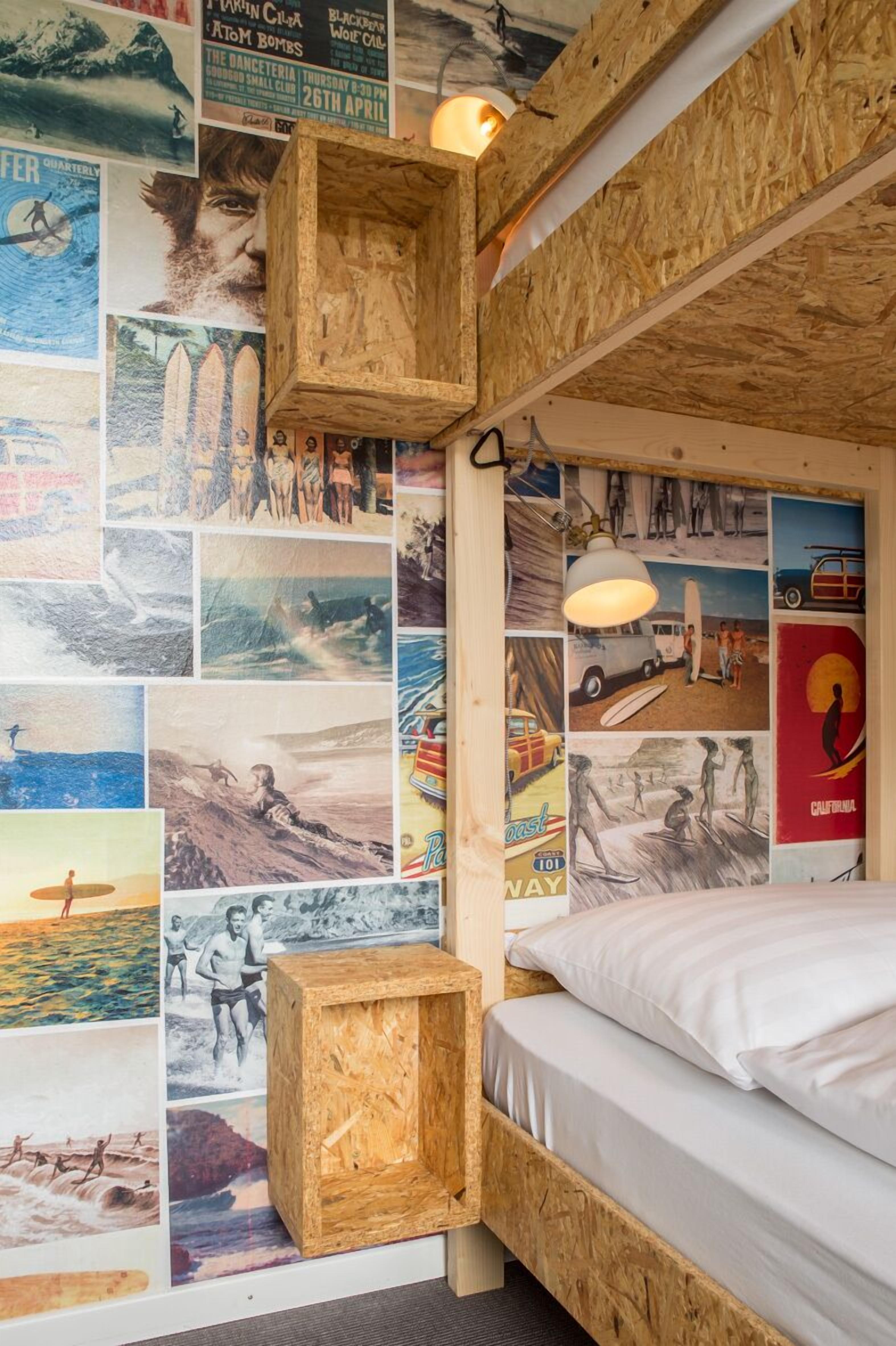 Quarto com cabeceira de madeira, cama com roupa de cama branca e parede decorada com diversos cartazes e fotografias.