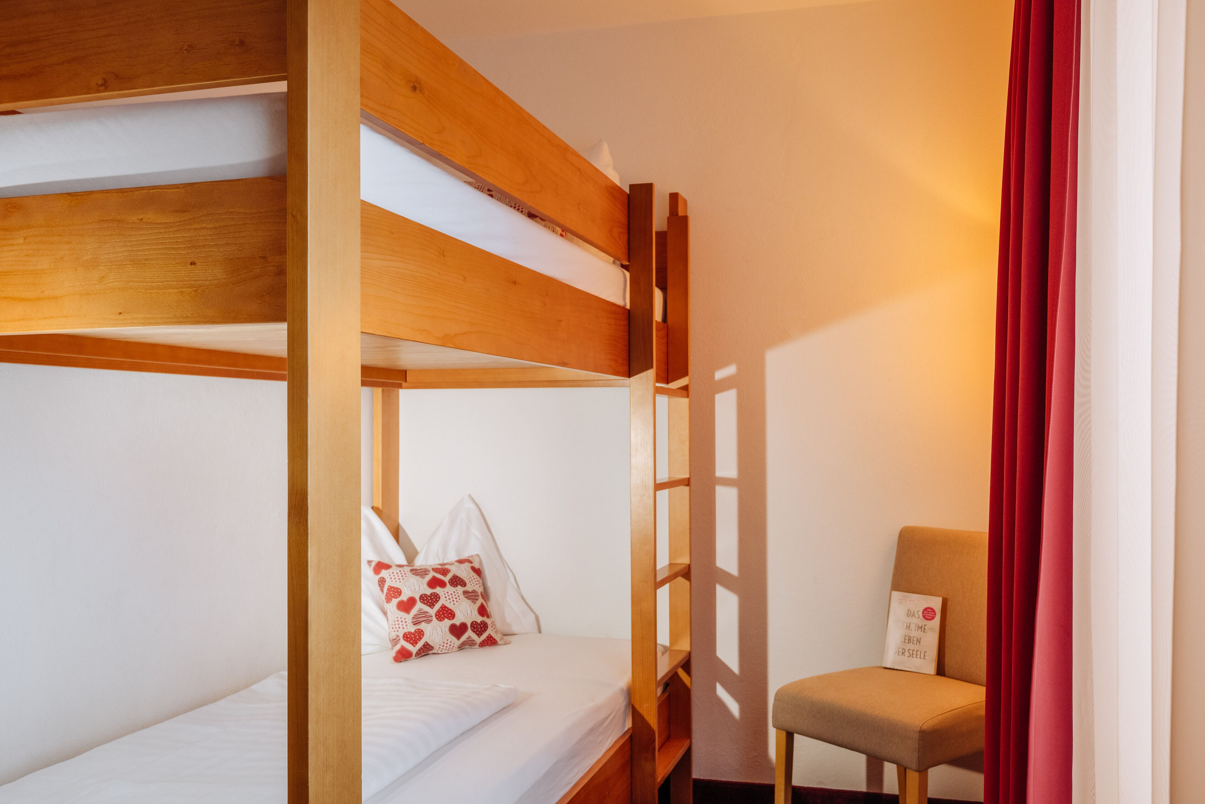 A bunk bed with a wooden frame, a single bed with white bedding and a heart-patterned pillow, and a chair with a cushion.