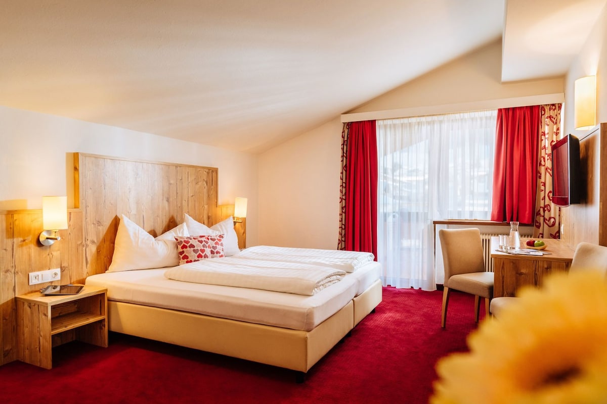 Quarto de hotel com cama de casal, mesas de cabeceira, escrivaninha com cadeira, televisão e janela com cortinas vermelhas.