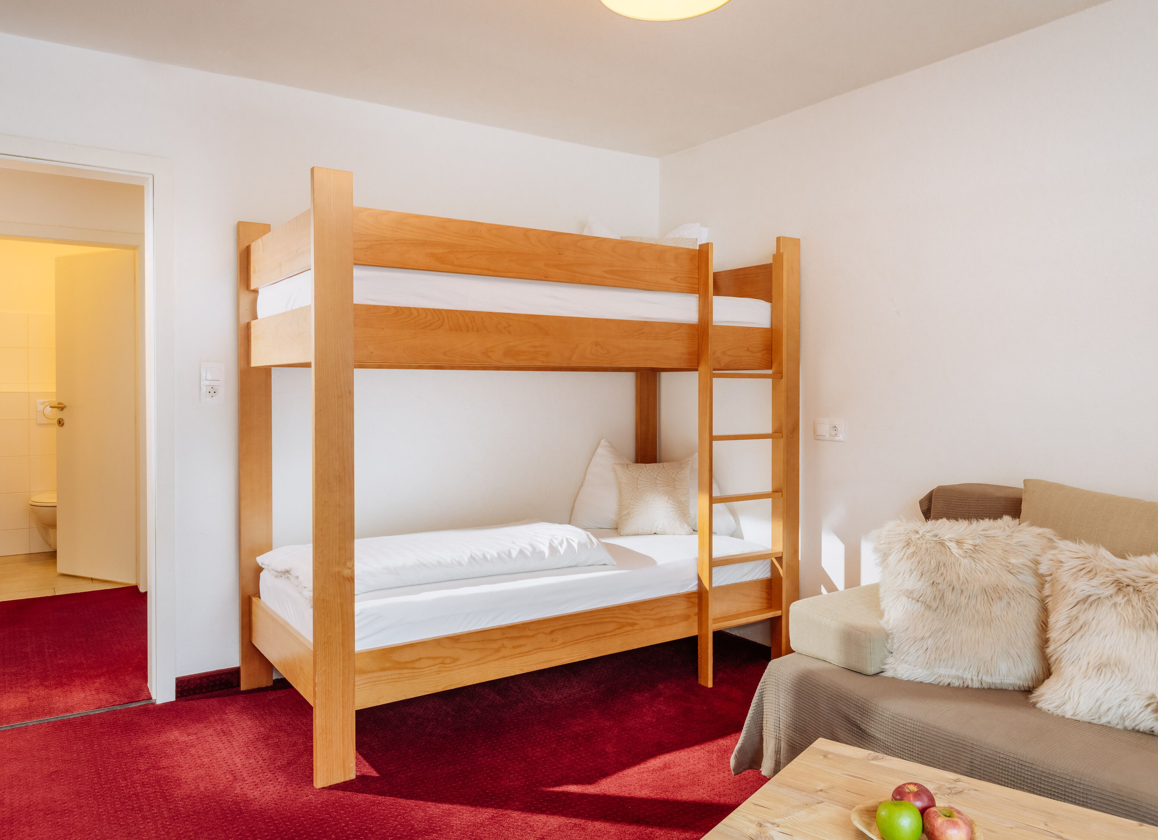 Una cama litera con estructura de madera, una alfombra roja, un sofá con una manta de pelo y una mesa con manzanas.