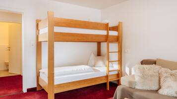 Una cama litera con estructura de madera, una alfombra roja, un sofá con una manta de pelo y una mesa con manzanas.