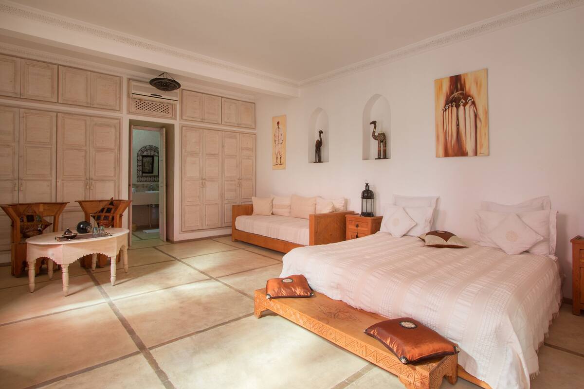 Een ruime slaapkamer met een groot bed, een houten bankje, een klein tafeltje en een garderobe.