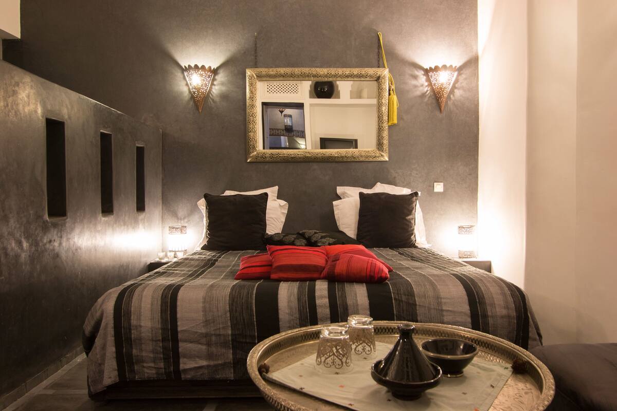 Een netjes opgemaakt bed met gestreept beddengoed en rode kussens, een nachtkastje met een dienblad met thee en glazen, en een wandspiegel met decoratieve verlichting.