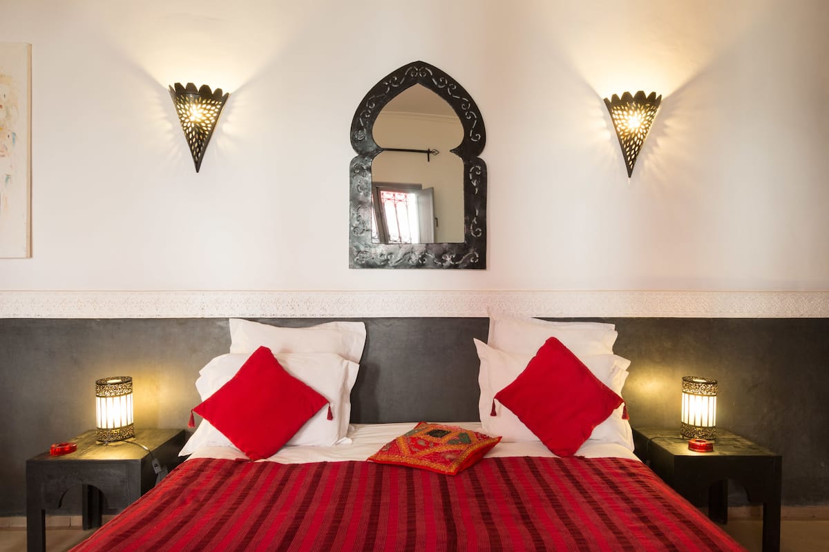 Een net opgemaakt bed met rood-witte beddengoed, twee bedlampjes en een spiegel aan de muur.