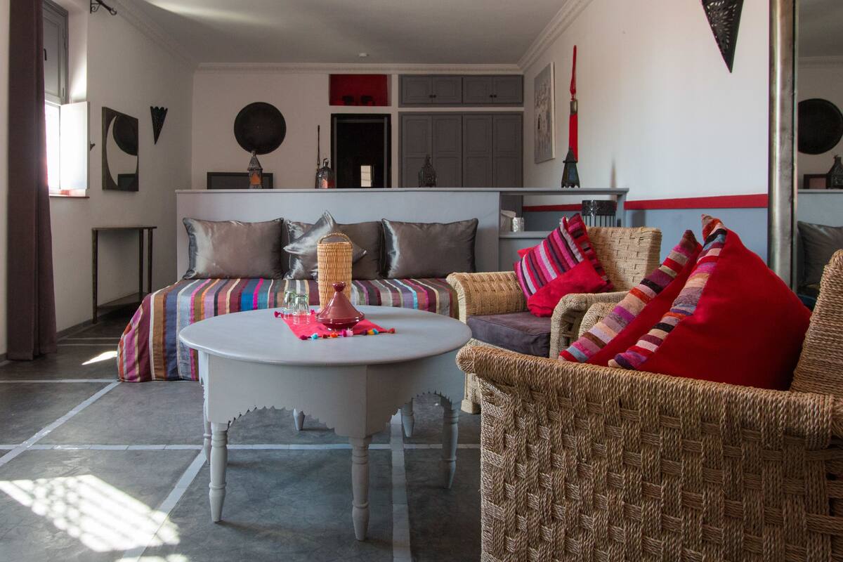 Een gezellige woonkamer met een ronde witte tafel, rieten stoelen en een kleurrijke gestreepte bank.