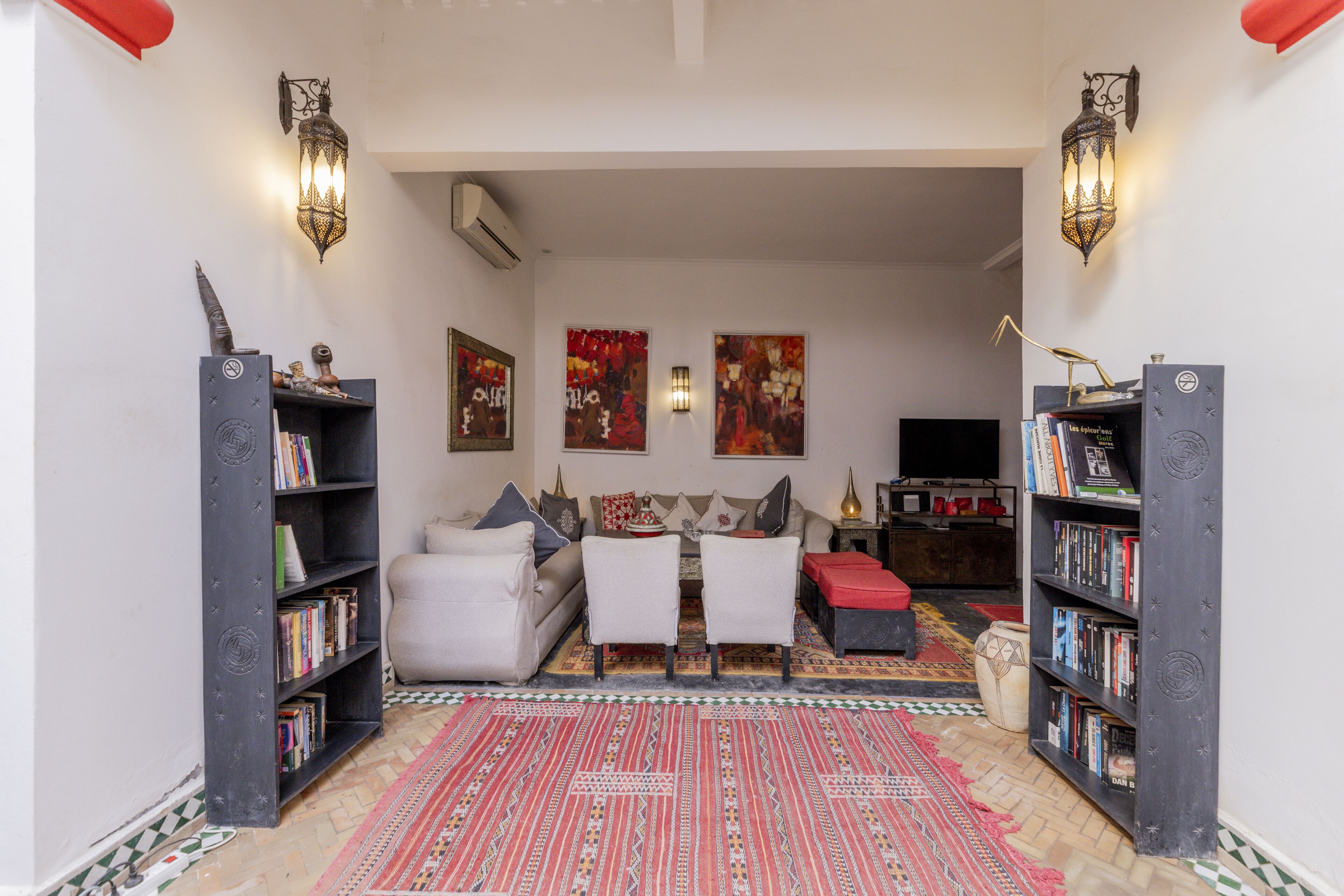 Ein Wohnzimmer mit einer Couch, zwei Sesseln, einem Couchtisch und einem Fernseher. An beiden Seiten befinden sich Bücherregale und die Wände sind mit Kunstwerken geschmückt.
