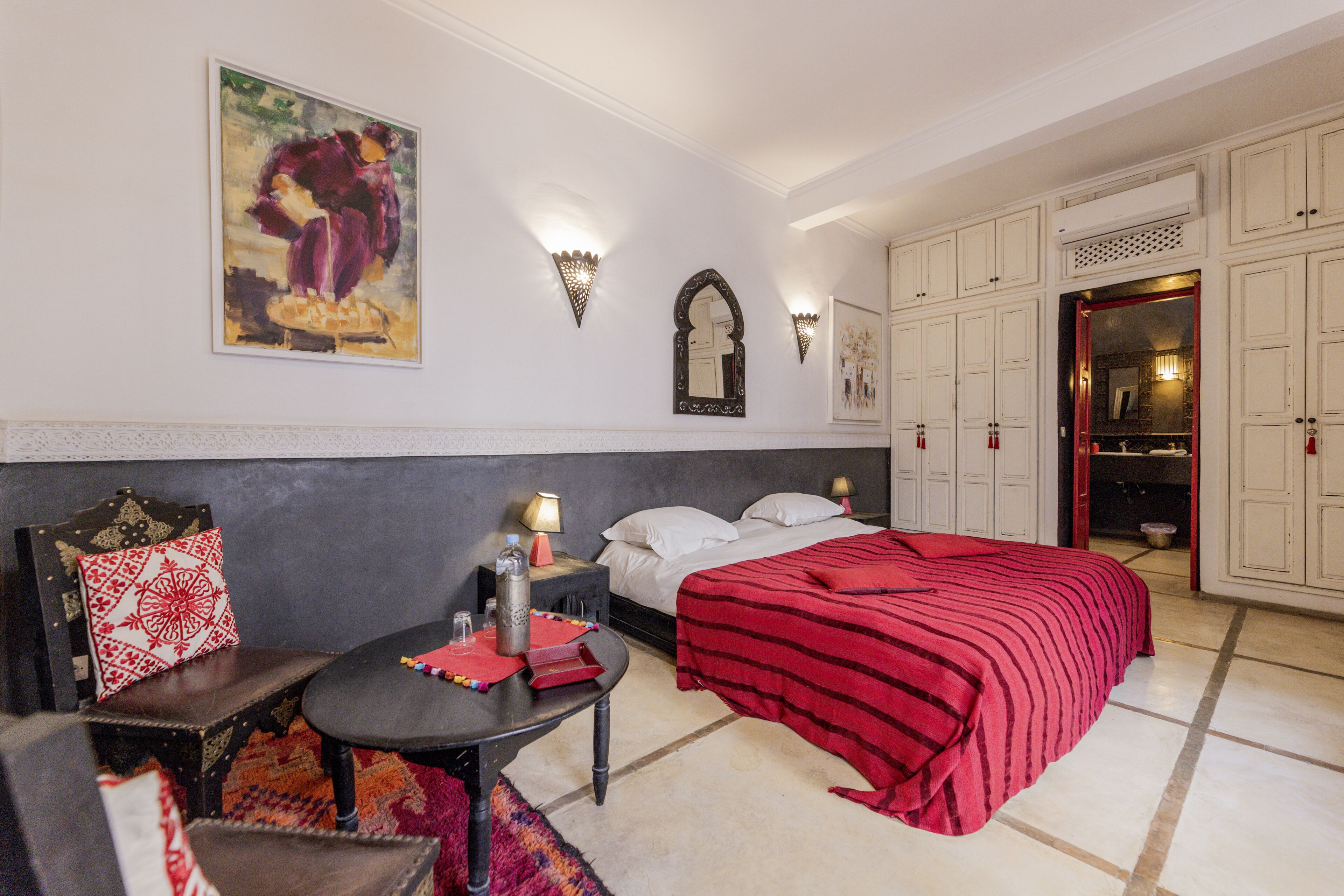 A hotel room with a bed, a table with a red cloth, a chair, a wall-mounted lamp, a painting, and a wardrobe.