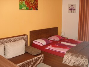 Habitación de hotel con cabecera de madera, cama con ropa de cama roja y rosa, mesita de noche con lámpara y una silla de mimbre.