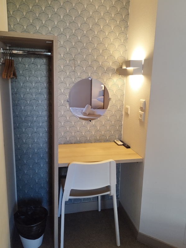 Una habitación pequeña con un espejo redondo, un escritorio de madera, una silla blanca y un cesto de basura.