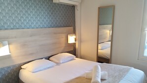 Una cama bien hecha con sábanas blancas, un plaid color beige y una toalla enrollada. Hay dos lámparas de noche y un espejo grande en la pared.