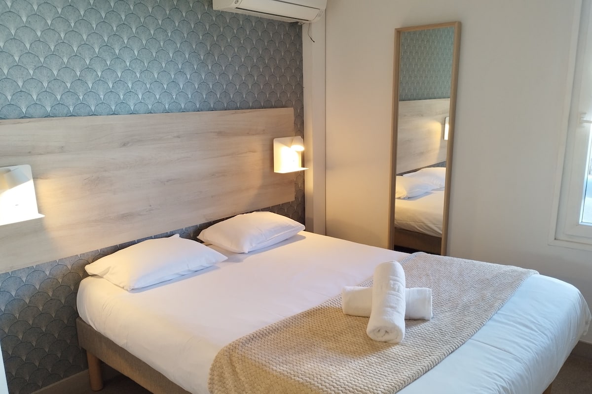 Una cama bien hecha con sábanas blancas, un plaid color beige y una toalla enrollada. Hay dos lámparas de noche y un espejo grande en la pared.