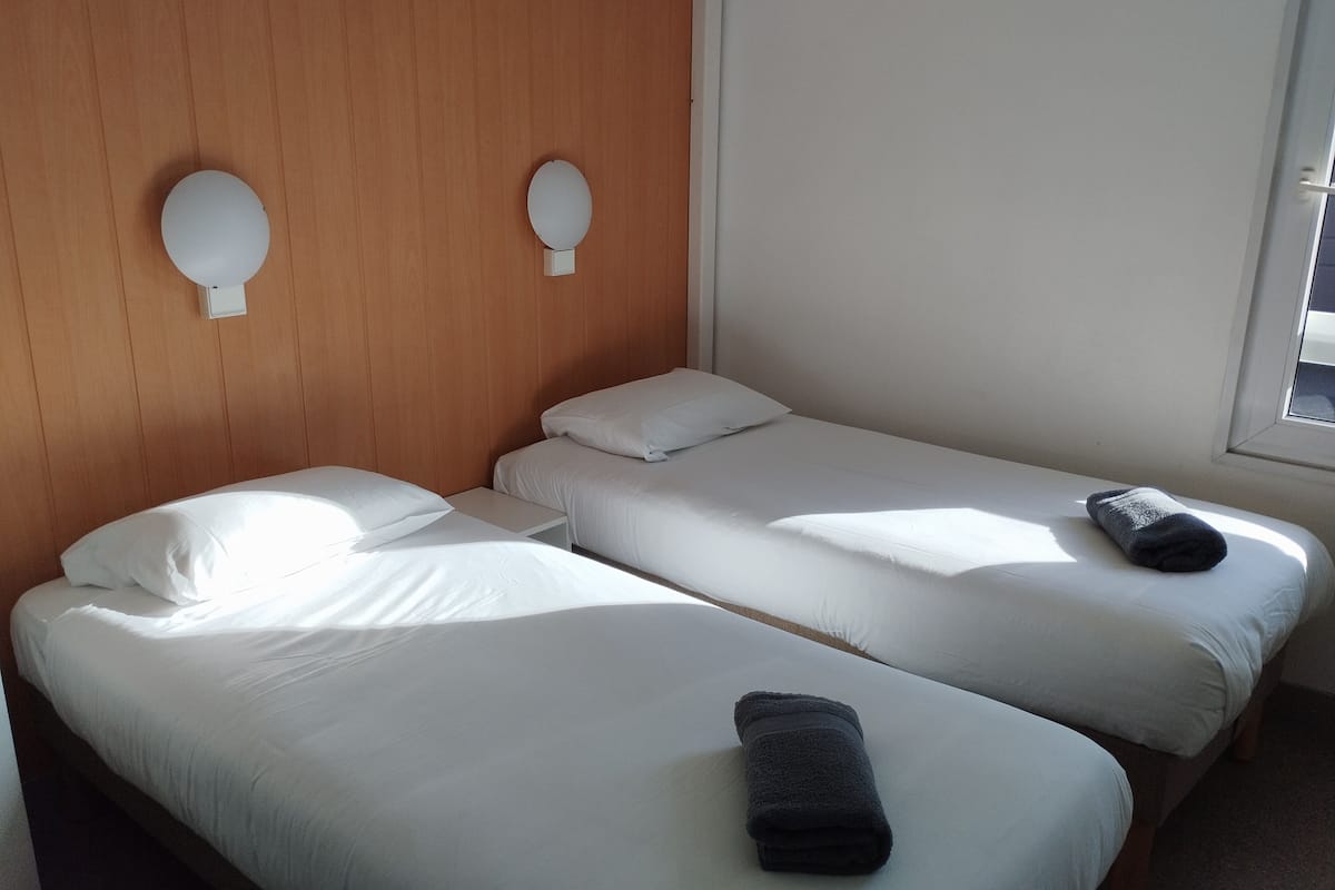 Dos camas individuales con ropa blanca de cama y una mesita de noche pequeña, un cabecero de madera y una ventana con luz natural.