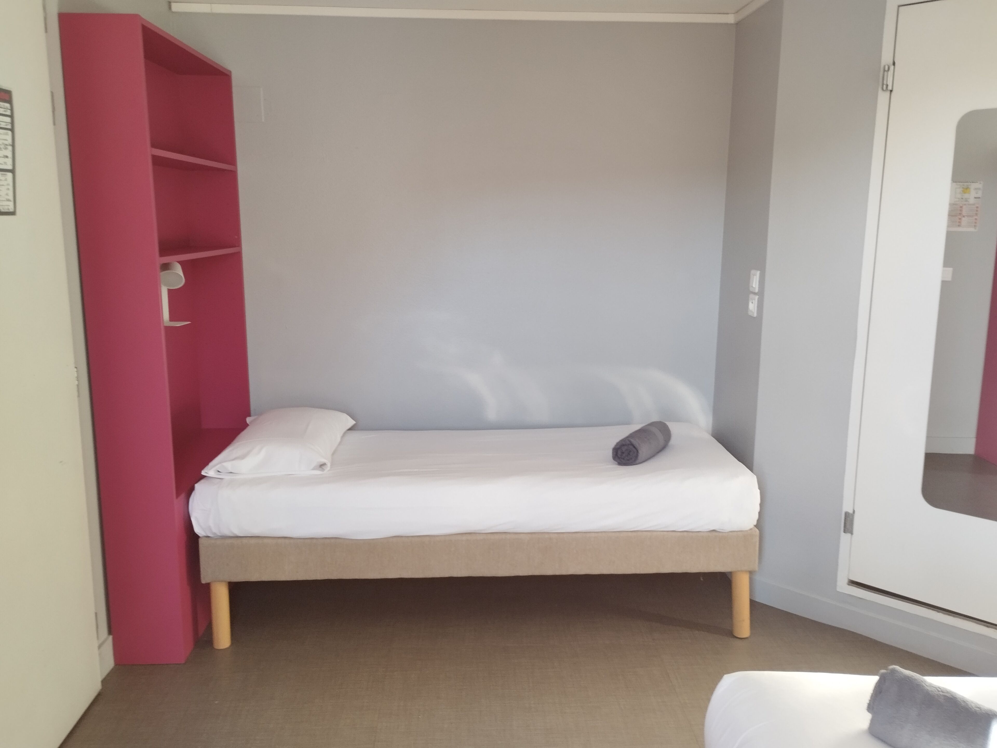 Quarto pequeno com uma cama de solteiro, guarda-roupa rosa, cama branca e porta branca.