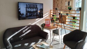 Una habitación moderna con un sofá marrón oscuro, una mesita con una revista, un trípode con una lámpara y un estante de madera con plantas.