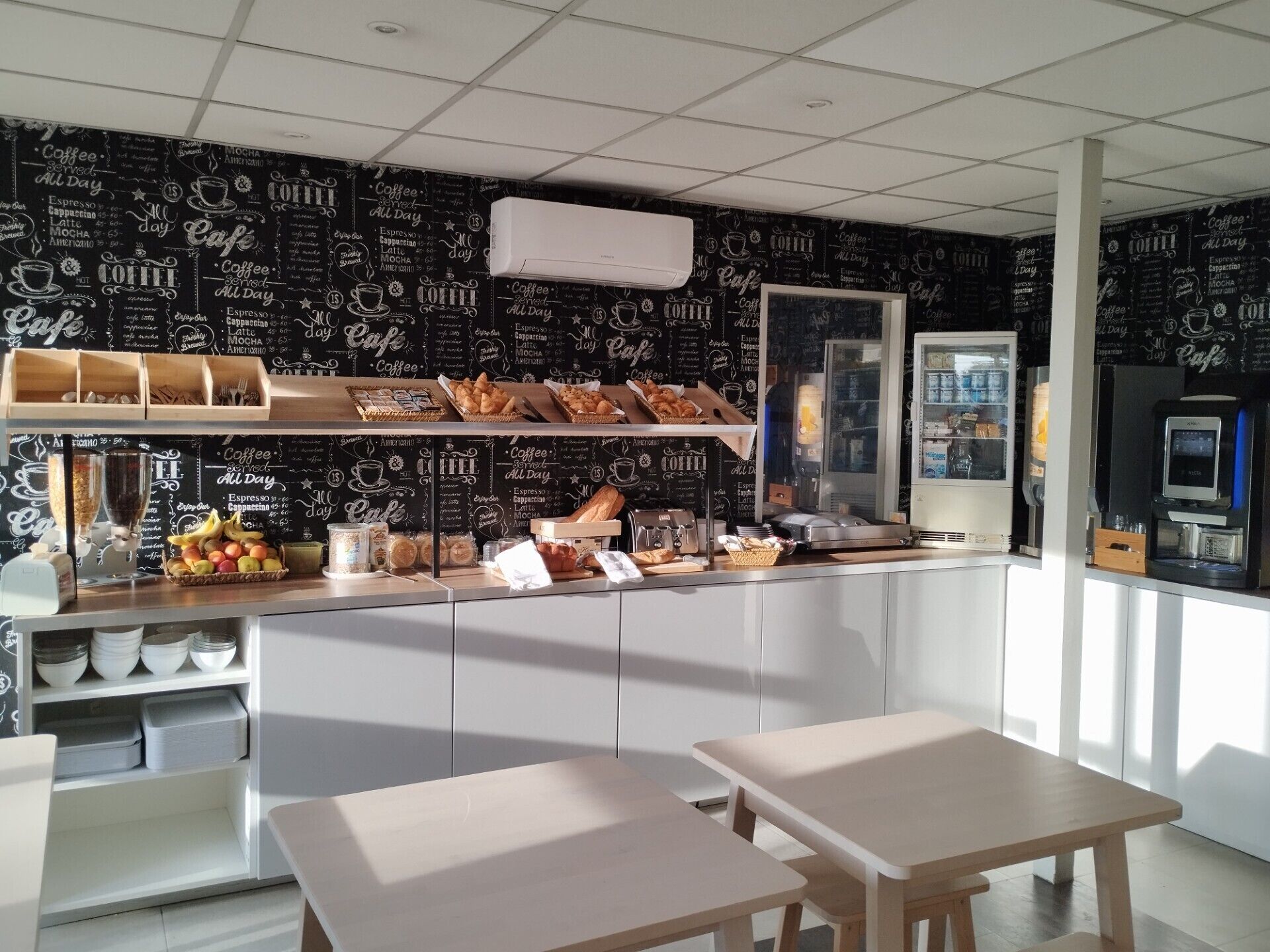 A café interior with a counter displaying pastries, a coffee machine, and a refrigerated display. There are two wooden tables and a white cabinet in the background.