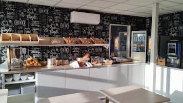 A café interior with a counter displaying pastries, a coffee machine, and a refrigerated display. There are two wooden tables and a white cabinet in the background.