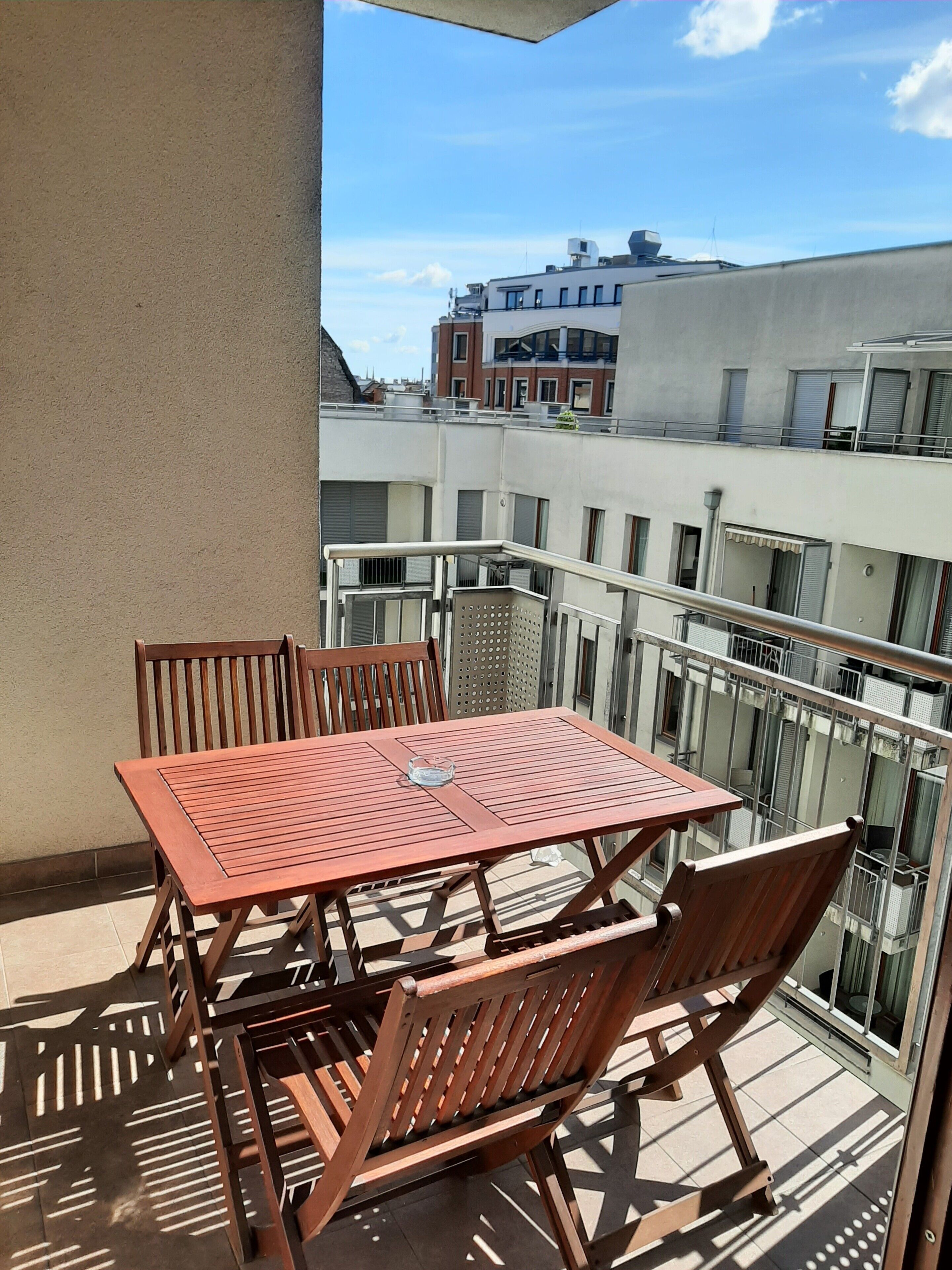 A balcony with a wooden table and chairs, a glass ashtray, and a view of surrounding buildings.