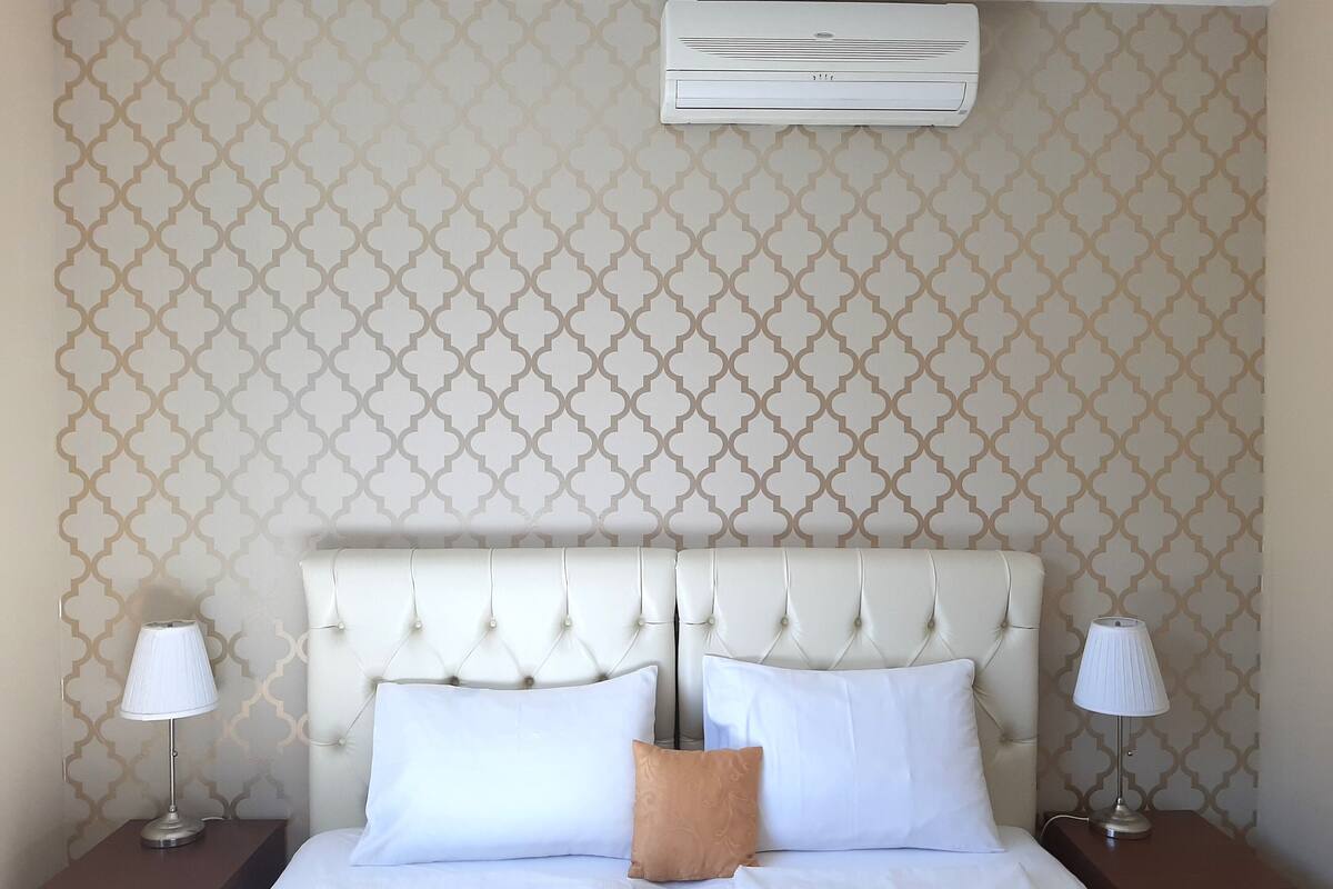 A hotel room with a tufted headboard, two bedside tables with lamps, a bed with white linens, and an air conditioning unit mounted on the wall.