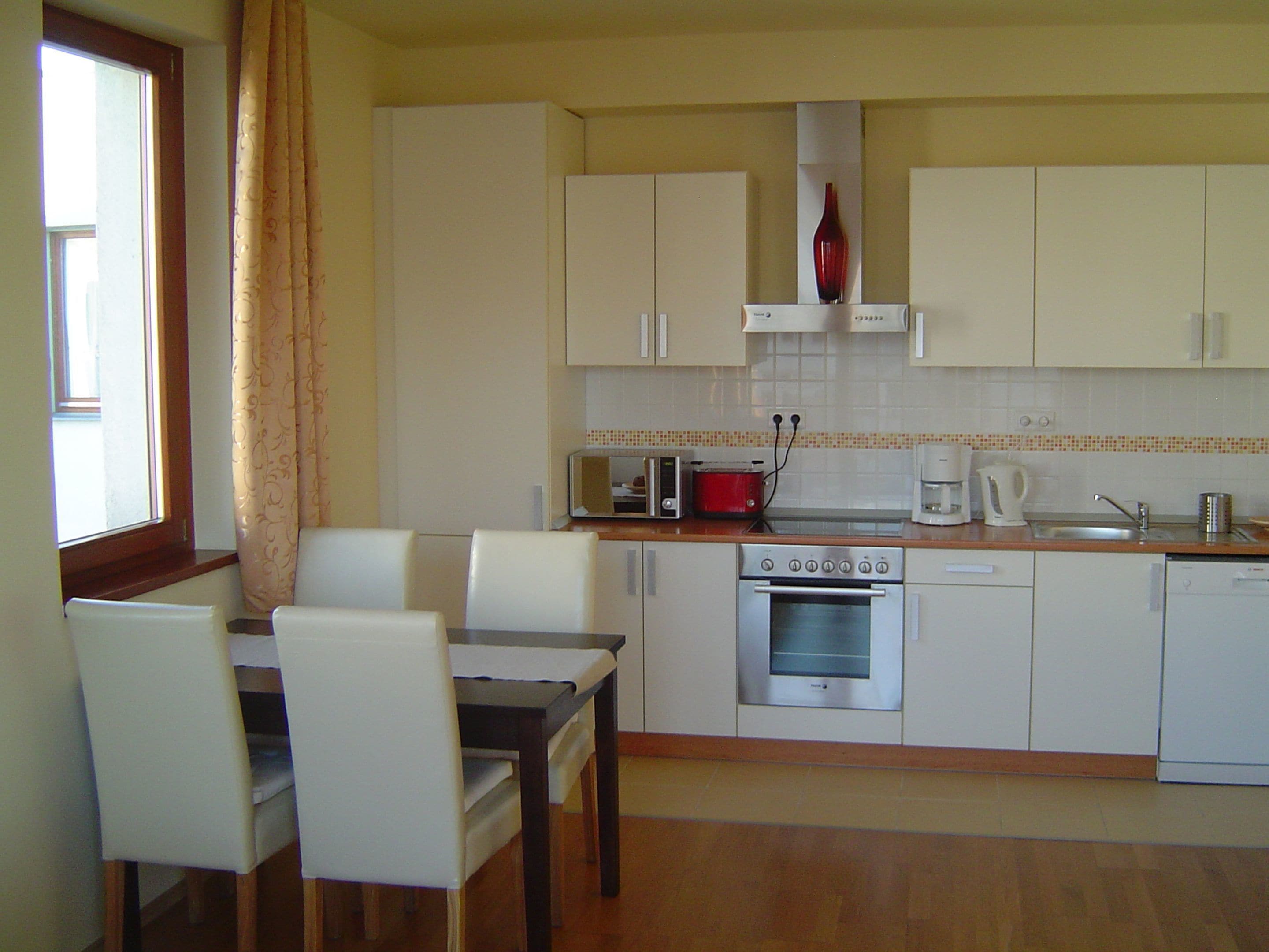 A compact kitchen with white cabinets, a dining table with chairs, a red toaster, a coffee maker, and a dishwasher.