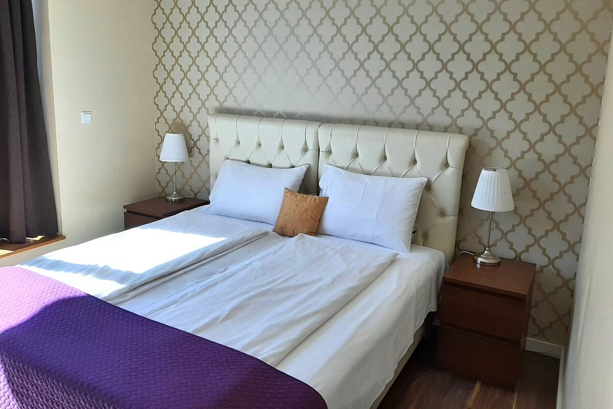 A neatly made bed with a headboard, two bedside tables with lamps, and a patterned wallpaper.