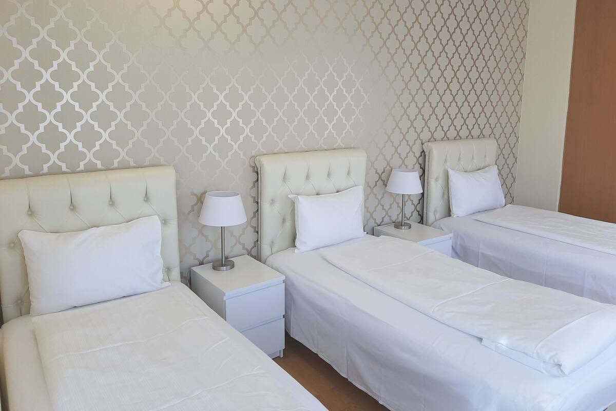 A hotel room with three beds, each with a white headboard and matching pillows. There are bedside tables with lamps and a patterned wall behind the beds.