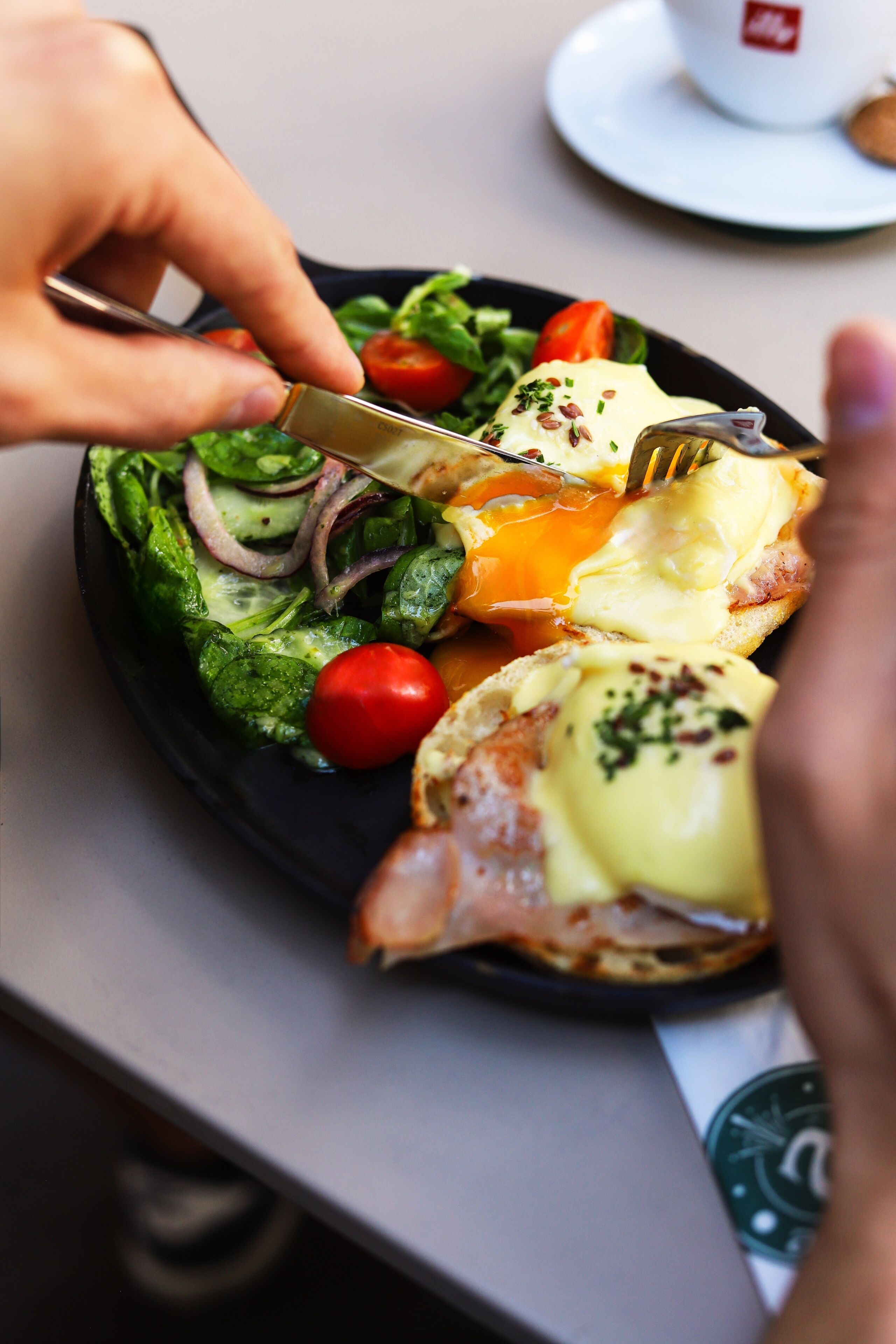 A person cutting an egg benedict on a plate with a side salad.