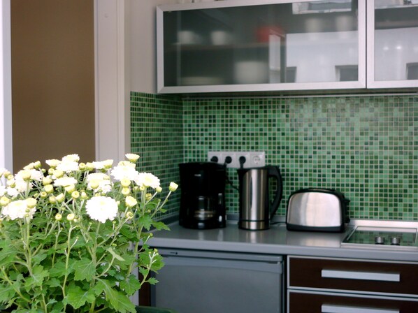 Une cuisine avec un mur recouvert de carreaux verts, une machine à café, un grille-pain et une plante en pot ornée de fleurs blanches.