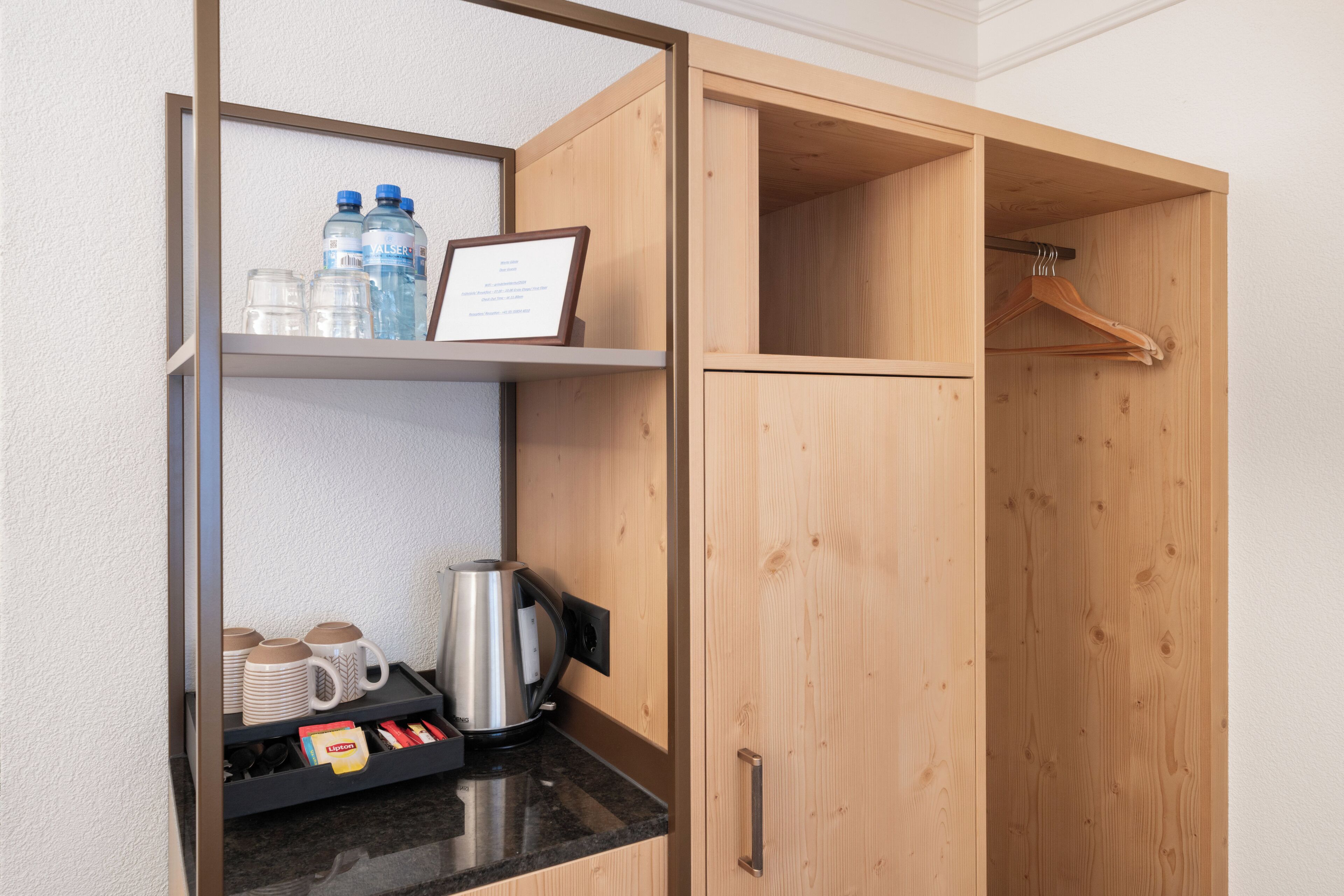 A wooden wardrobe with a hanging space, a shelf with water bottles, mugs, and a framed certificate.