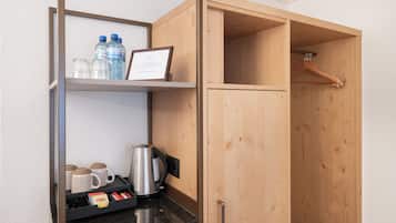A wooden wardrobe with a hanging space, a shelf with water bottles, mugs, and a framed certificate.