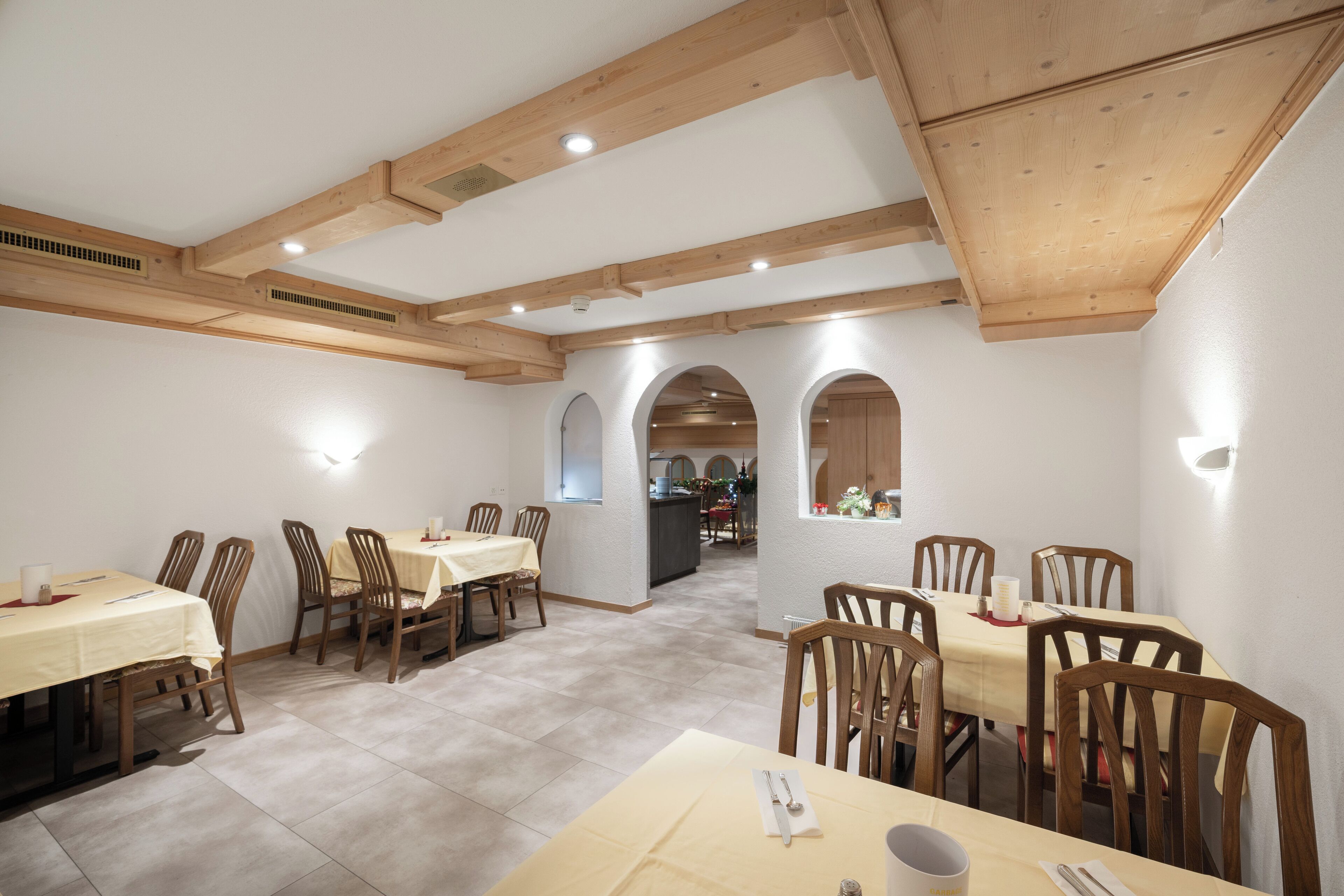 A dining area with wooden tables and chairs, white walls, and a kitchen in the background.
