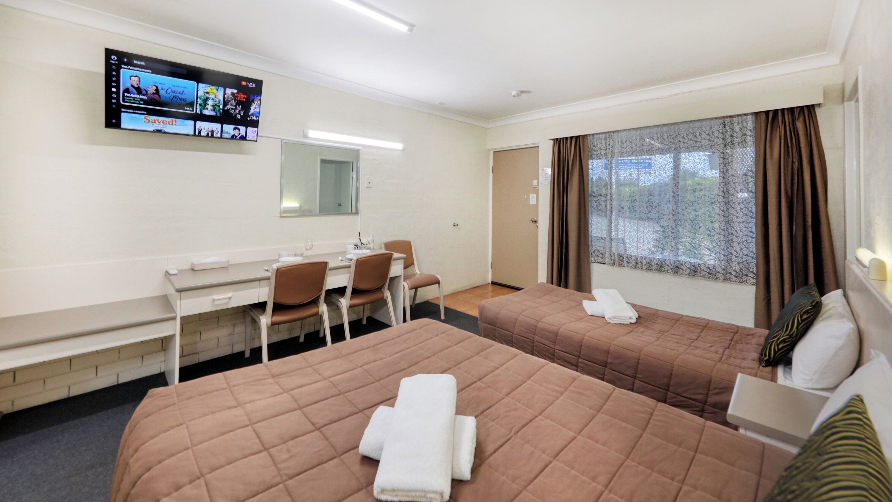 A hotel room with two beds, a desk with chairs, a TV mounted on the wall, and a window with curtains.