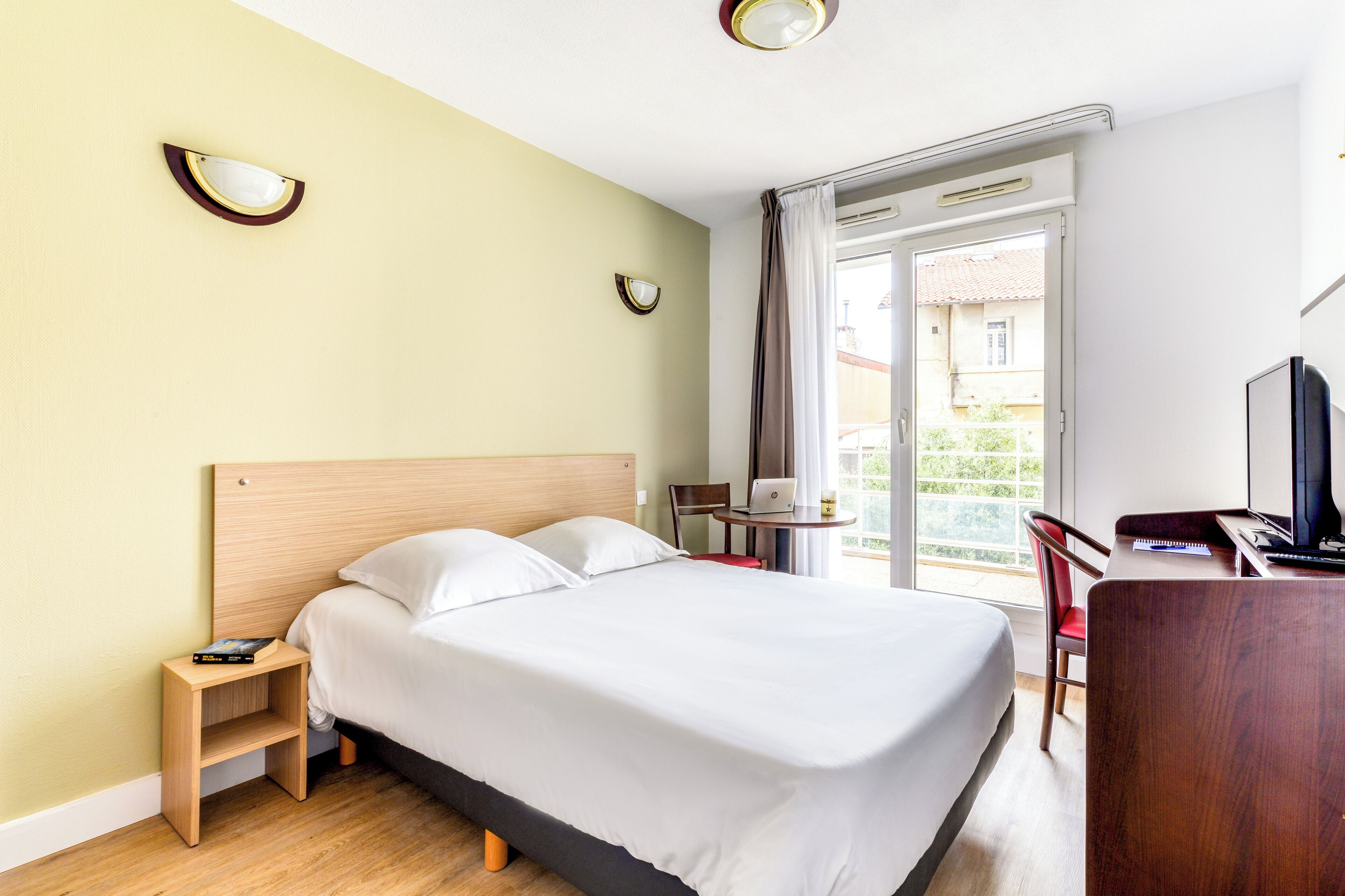 A hotel room with a bed, bedside table, desk, chair, and a view of the outside.