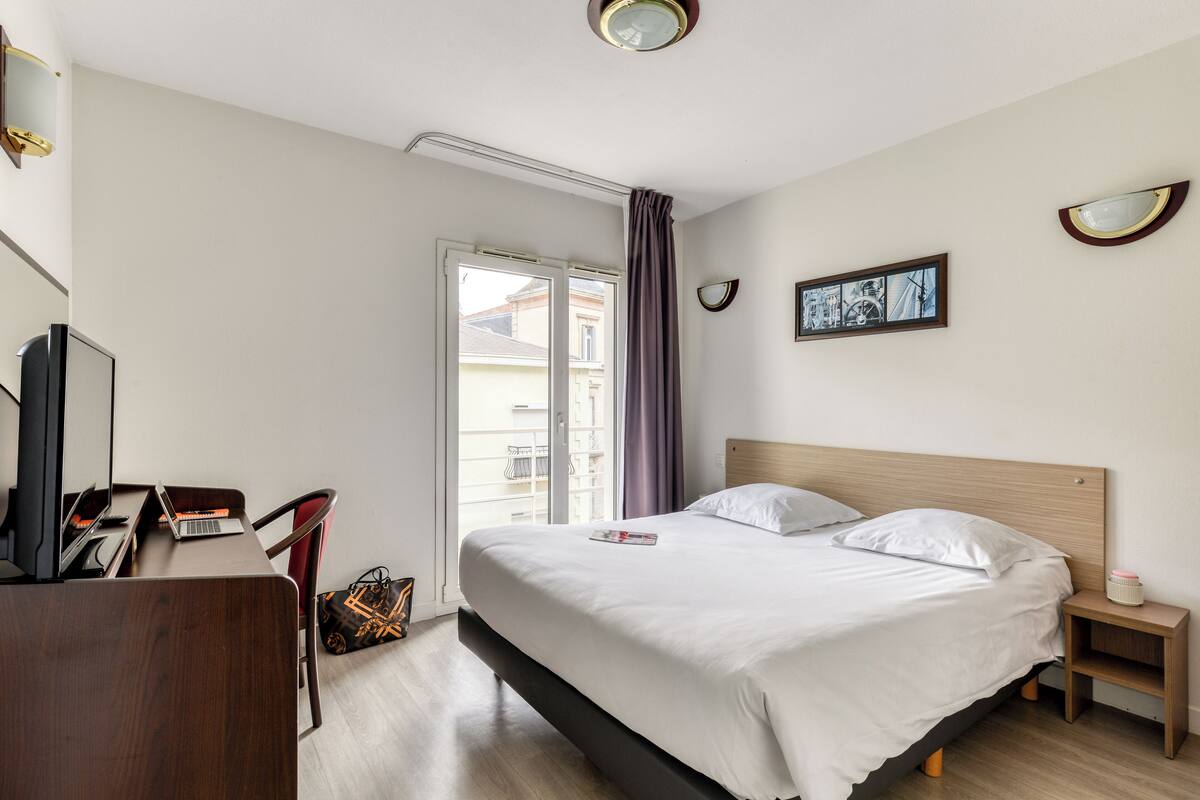 A hotel room with a bed, a desk with a laptop, a chair, a small bedside table with a lamp, and a window with curtains.