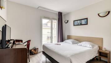 A hotel room with a bed, a desk with a laptop, a chair, a small bedside table with a lamp, and a window with curtains.