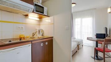 A compact hotel room with a kitchenette, a bed, a desk with a laptop, and a window with curtains.