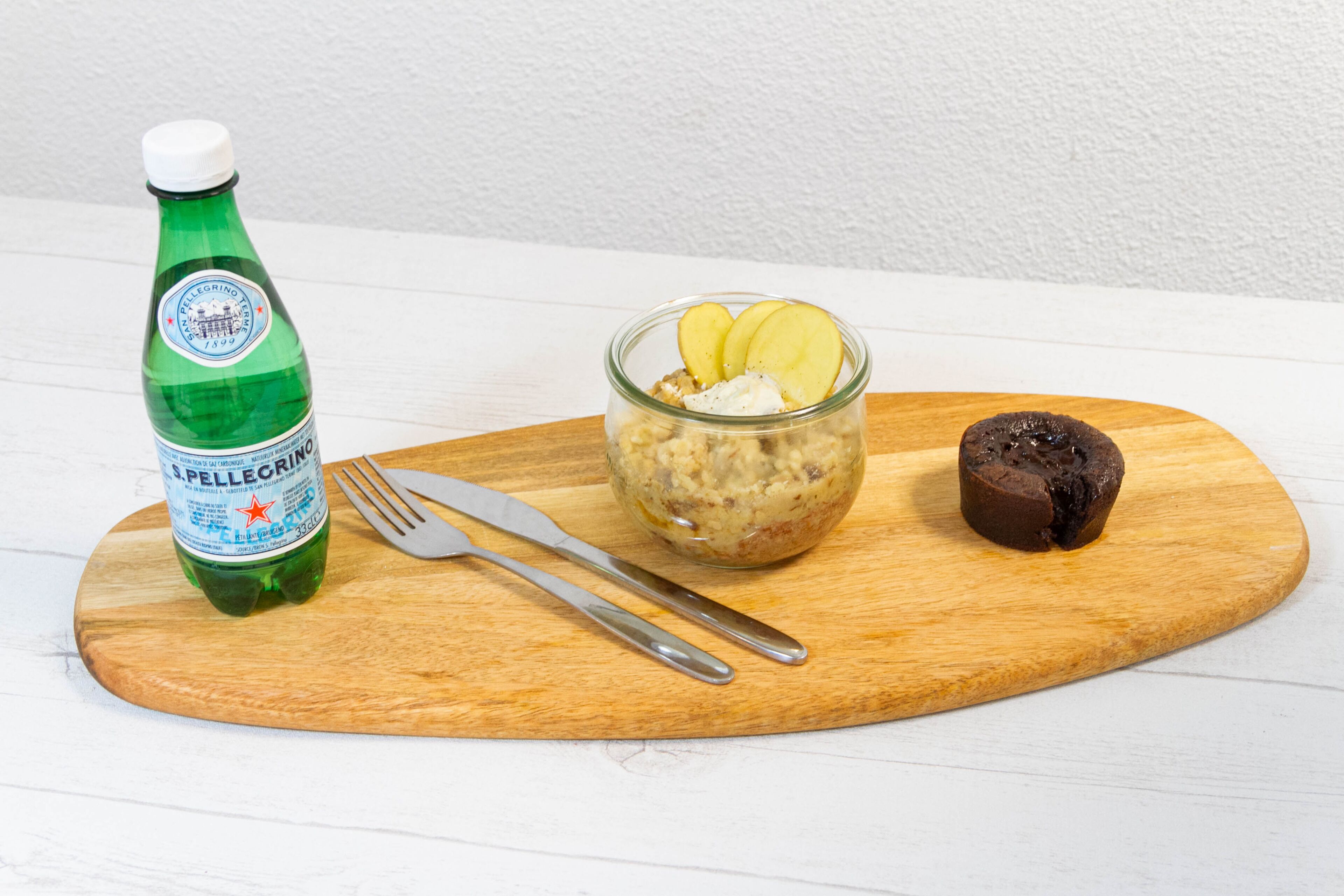 Pullo S. Pellegrino -mineraalivettä, lasikulho, jossa on jälkiruokaa, ja pala suklaata puulautalla.