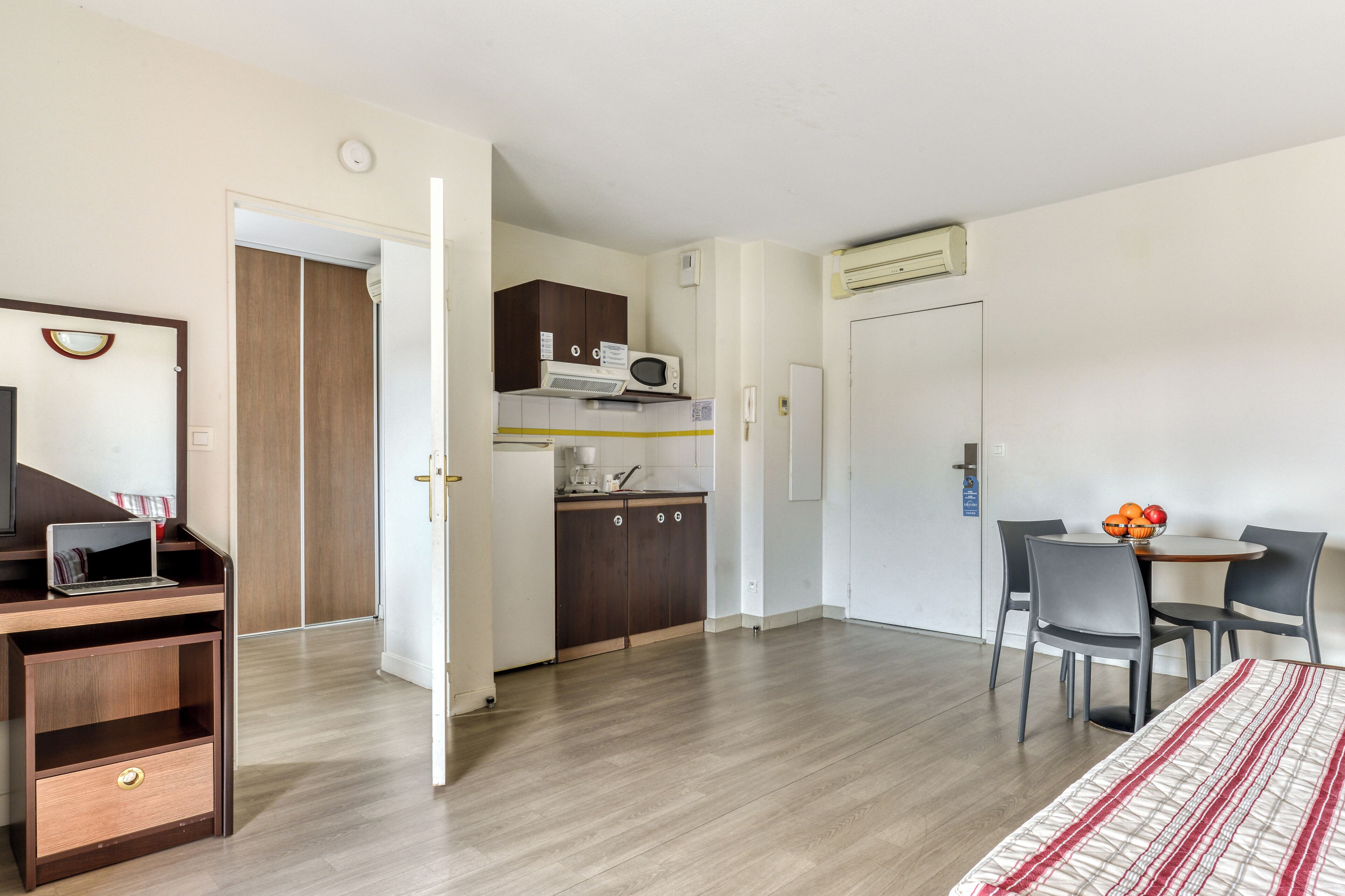 A compact hotel room with a wooden desk, a small kitchenette, a dining area with a table and chairs, and a bed with a striped bedspread.